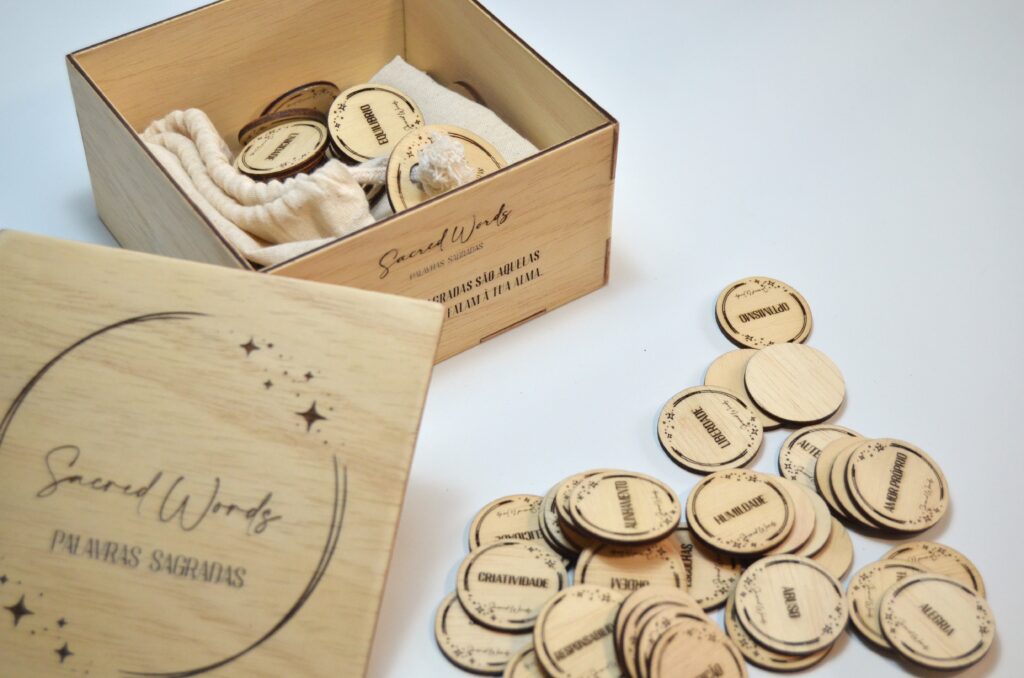 Kit Sacred Words com caixa de madeira aberta, peças redondas com palavras de poder (Transceder, Vida, Otimismo) e um saco de tecido para guardar as peças.